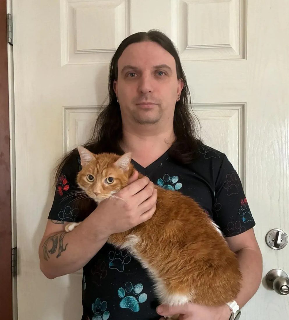 Man with long hair holding an orange cat in front of a white door