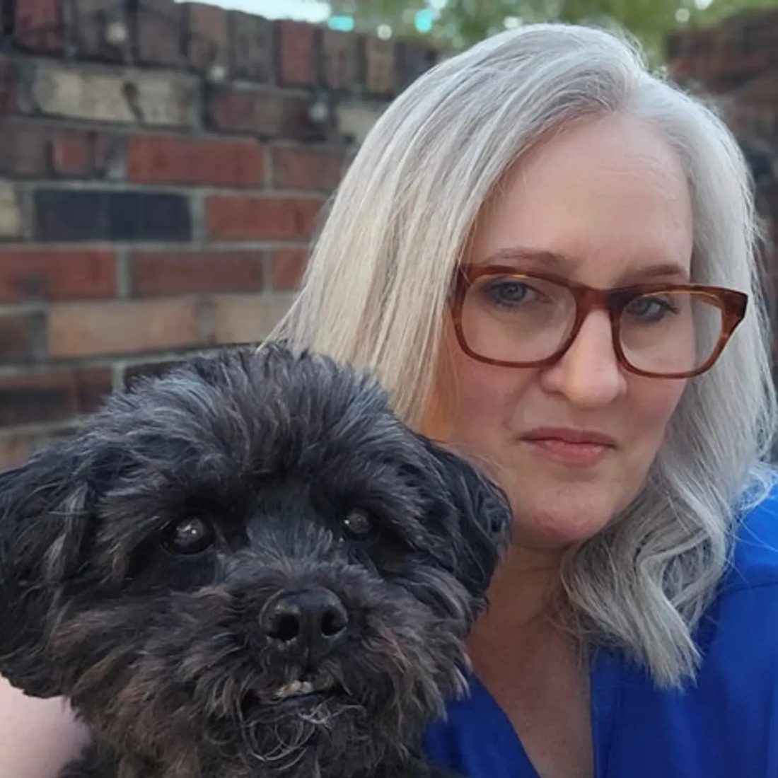 Woman with glasses holding a small black dog in front of a brick wall